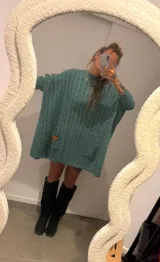 Sweater de punto color camel con diseño de trenzas verticales y acabado brillante. Presenta una silueta holgada tipo poncho, cuello redondo, mangas largas con puños ajustados y bolsillos frontales con detalle de etiqueta de cuero.