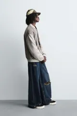 Cazadora estilo varsity o bomber de hombre, color gris oscuro con efecto ante, mangas en tono gris claro y puños, cuello y bajo con rib elástico a rayas blancas y grises. Presenta un parche con la letra 'R' en el pecho derecho y un bordado con texto en el pecho izquierdo.