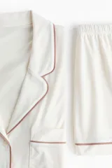 Pijama de dos piezas confeccionado en mezcla de viscosa, compuesto por una camisa de manga corta con cuello abierto, botones frontales y bolsillo superior, y un short con elástico en la cintura. Ambos presentan ribetes decorativos en contraste.