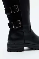 Bota alta de cuero sintético negro con suela track y tacón bajo. Presenta detalle de cuatro correas con hebillas metálicas en el lateral exterior de la caña.