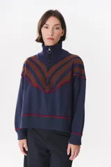 Buzo de tejido de punto en color azul marino, con cuello alto y cierre frontal parcial. Presenta un diseño con franjas en color marrón y detalles de líneas en rojo en el pecho, hombros y ruedo. Tiene corte holgado y mangas largas con puños acanalados.