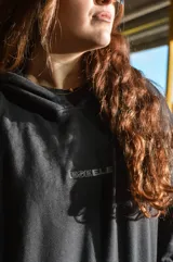 Canguro negro con capucha, corte oversize y diseño con hombros caídos. Presenta detalles bordados en la espalda con la palabra "Dual" en tipografía cursiva junto a texto descriptivo en tonos monocromáticos.