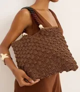Bolso de mano tipo tote, tejido en punto de ganchillo con textura de conchas, color marrón oscuro. Las asas son de cuero o material sintético oscuro, adornadas con cuentas redondas de madera en tonos marrón oscuro, borgoña y crema.