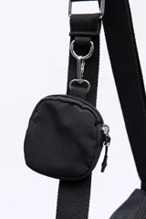 Cartera tipo bandolera de nylon color negro, con correa ajustable y detalle de hebilla metálica lateral. Presenta un diseño minimalista con cierre superior y logo sutil en la parte inferior.