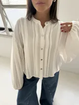 Blusa blanca con cuello mao y cierre frontal de botones de nácar. Presenta mangas largas abullonadas con puños elásticos y detalle de tablas verticales en el frente.