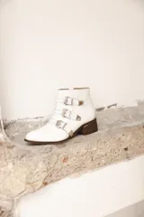 Botineta blanca de cuero, con taco cuadrado de madera, punta afilada y tres tiras con hebillas metálicas y tachas decorativas.