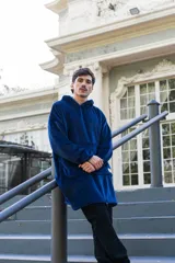 Buzo tipo manta con capucha, confeccionado en tela de peluche suave color azul marino. Presenta un diseño versátil con cordón ajustable en el ruedo para modificar el largo, capucha amplia y un bolsillo frontal de gran tamaño.