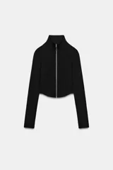 Sudadera crop entallada de color negro, con cuello alto, manga larga y cierre frontal de cremallera metálica. Presenta un diseño corto con bajo asimétrico.