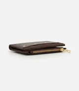 Billetera mediana color marrón, confeccionada en poliuretano con textura lisa y detalle de gamuza en la parte superior. Presenta cierre superior con cremallera dorada y aplicación de tachas metálicas decorativas en el frente.