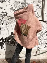 Campera tipo abrigo de textura borreguito en color rosa, con diseño de collage en la espalda que incorpora un parche de pana rojiza y un panel central con estampado geométrico étnico. Presenta capucha integrada y un detalle de etiqueta de cuero sintético en la parte posterior.