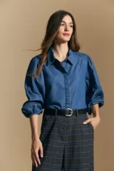 Camisa de denim azul con cuello camisero, cierre frontal de botones y mangas largas abullonadas con hombros fruncidos.