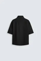 Sobrecamisa negra de tejido con elasticidad, cuello solapa y manga corta. Tiene dos bolsillos de plastrón en el pecho y cierre frontal con botones a presión.