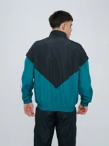 Campera deportiva Umbro modelo Beat, confeccionada en nylon ligero con diseño de bloques de color en blanco, azul marino y verde azulado. Presenta cierre frontal, cuello alto y elásticos en puños y cintura.