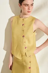 Top de lino blanco sin mangas, con cuello redondo y cierre de botones de madera dispuestos en una cartera asimétrica. Cuenta con delicados detalles bordados en forma de flor alrededor de los ojales.