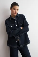 Camisa de jean azul oscuro con cierre frontal de botones metálicos a presión y dos bolsillos delanteros con solapa en el pecho. Presenta mangas largas con puños anchos doblados y cuello camisero clásico.