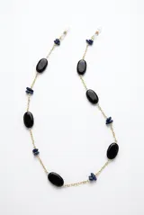 Correa para lentes con cadena dorada, intercalada con cuentas ovaladas negras y pequeñas piedras azules. Cuenta con sujetadores de silicona en los extremos.