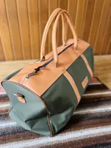 Bolso de viaje tipo duffel de lona negra con detalles en cuero tostado, asas de mano y correa ajustable para el hombro.