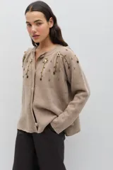 Saco de tejido de punto color beige, con diseño de cuello redondo y cierre frontal con botones forrados en crochet. Presenta un delicado bordado floral artesanal que cae desde los hombros, con mangas largas y corte clásico.