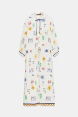 Vestido largo estilo túnica de color crudo con estampado multicolor de motivos marinos y mediterráneos. Presenta cuello con volante, cierre frontal con botones, manga larga y cordón ajustable en el cuello con abalorios.