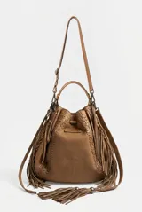 Cartera estilo saco de cuero vacuno marrón, con flecos colgantes, asa tubular rígida y detalles de costuras decorativas. Cuenta con cordón ajustable en la parte superior adornado con borlas y herrajes metálicos.