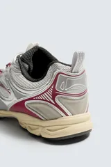 Zapatilla deportiva tipo running con diseño técnico, confeccionada en una combinación de materiales sintéticos y malla. Presenta una estética metalizada en tonos plata con detalles en color granate y una suela con volumen de diseño irregular en tono crema.