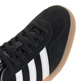 Championes Adidas Gazelle Indoor de diseño clásico, confeccionados en gamuza negra con las icónicas tres tiras laterales en blanco. Presentan el nombre del modelo en letras doradas, talón en contraste blanco y suela de caucho color caramelo.