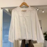Camisa blanca de manga larga con detalles de encaje y bordados en los hombros y el frente, cuello en V con volados y cierre frontal con botones.