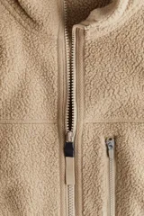 Campera deportiva color beige en tejido sherpa, con cuello alto, cierre frontal, bolsillo superior con cierre, bolsillos laterales con cierre, puños y bajo con elástico fino.