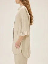 Camisa de lino de silueta relajada y largo irregular, color beige, con cuello mao, botones frontales a contratono y mangas tres cuartos terminadas en volados.