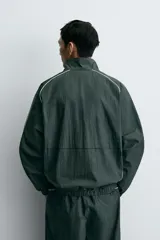 Chaqueta técnica de corte relajado en color verde oscuro, con cuello subido, cierre frontal de cremallera y detalles de líneas en contraste. Presenta bolsillos frontales con cremallera oculta y acabados elásticos en puños y ruedo.