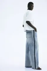 Jean de tiro alto, confeccionado en denim de algodón rígido, de corte ancho y pierna recta. Presenta un lavado celeste claro con efecto desgastado en el centro y franjas laterales en denim más oscuro con acabado desflecado.