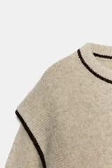 Jersey de punto color beige con cuello redondo y mangas largas abullonadas. Presenta detalles de vivos en color negro en el cuello, hombros y puños.