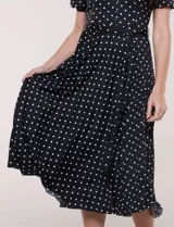 Vestido midi negro con estampado de lunares blancos, de la marca Sharagano. Presenta cuello con solapa, escote en V, cierre frontal con botones y mangas cortas. Tiene un corte en la cintura con lazo ajustable y falda plisada.