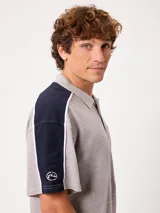 Remera polo gris melange de ajuste holgado, con cuello y cierre frontal de botones. Presenta detalles en azul marino y blanco en los hombros y mangas, y el logo de la marca bordado en el pecho.