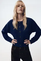 Chaqueta de punto de corte cropped y manga larga, con cuello redondo y cierre frontal de botones. Presenta dos bolsillos de plastrón en la parte delantera y acabados en rib.