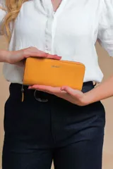 Billetera larga tipo sobre de la marca Lincoln's, confeccionada en material sintético con textura suave en color beige. Presenta un cierre perimetral metálico y el logo de la marca grabado en la parte frontal.