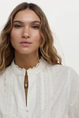 Camisa blanca de algodón con cuello alto, detalles de encaje broderie en el frente, mangas abullonadas y puños con volados y encaje. Cierre frontal con botones tapados.