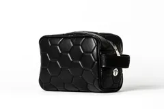 Necessaire de viaje o neceser de mano, color negro, con diseño acolchado en forma de hexágonos que simulan la textura de un balón de fútbol. Posee cierre de cremallera y una correa lateral de tela.
