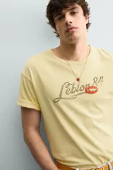 Camiseta de corte relajado color amarillo con efecto lavado. Presenta cuello redondo, mangas cortas con dobladillo y un estampado frontal con texto y un parche decorativo.