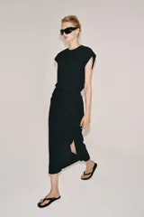Vestido midi negro con cuello redondo y manga sisa, confeccionado por Zara Woman Collection. Presenta un diseño drapeado en la parte delantera y un bajo asimétrico con un voluminoso volante lateral. Incluye un pequeño cierre de botón en la espalda.