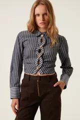 Camisa corta a rayas verticales grises y blancas, con cuello clásico y mangas largas. Presenta un diseño trenzado en el frente con botones dorados y aberturas.