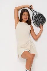 Vestido corto deportivo color beige, sin mangas y con cuello redondo. Presenta un diseño tipo pollera envolvente con lazo lateral ajustable en la cintura y logo pequeño estampado en el pecho.