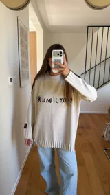 Sweater oversized crudo de tejido de punto, con hombros caídos y mangas largas. Presenta líneas negras en contraste en los hombros, bordado artesanal con la frase "e pur si muove" en el frente y aberturas laterales en el dobladillo.