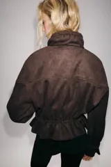 Cazadora marrón de efecto ante con cuello subido, manga larga con puños ajustados y bolsillos frontales. Cuenta con cierre frontal de cremallera y botones ocultos, además de detalles de trabillas con botones dorados en los laterales y puños.