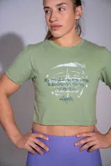Remera crop top de algodón elastizado color verde militar, con mangas cortas, cuello redondo y estampado frontal con texto y diseño gráfico en tonos blancos y oscuros.