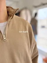 Buzo gris oscuro de corte oversize con cuello alto y cierre frontal parcial. Cuenta con mangas largas y puños elásticos, además de un pequeño bordado con texto en el pecho.