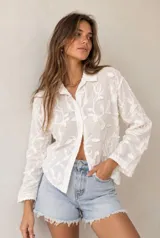 Camisa blanca de manga larga con diseño bordado floral en relieve, cuello clásico y cierre frontal de botones.
