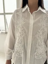 Camisa blanca de manga larga con bordados florales al tono en el frente, cuello clásico y cierre frontal de botones.