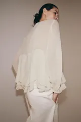 Blusa de viscosa color rosado con cuello redondo, manga larga abullonada con puños elásticos y cierre frontal con lazadas. Presenta un diseño con volantes en el bajo y corte asimétrico.
