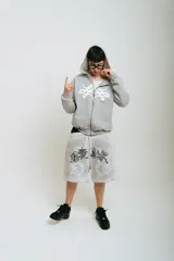 Bermuda de felpa color gris claro con efecto lavado (washed). Presenta un corte ancho con cintura elástica y cordón beige. El diseño frontal incluye un estampado serigrafiado en negro con texto y motivos florales/ornamentales en bordado blanco. Posee remaches metálicos cromados en los laterales y bolsillos.